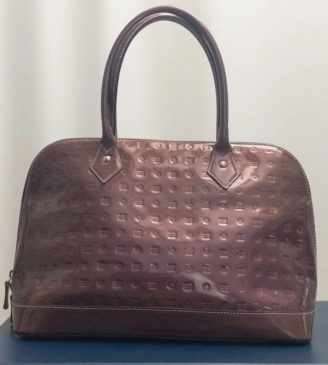 A brown textured leather handbag with a domed shape, double top handles, and a glossy finish, displayed on a blue surface against a light background.