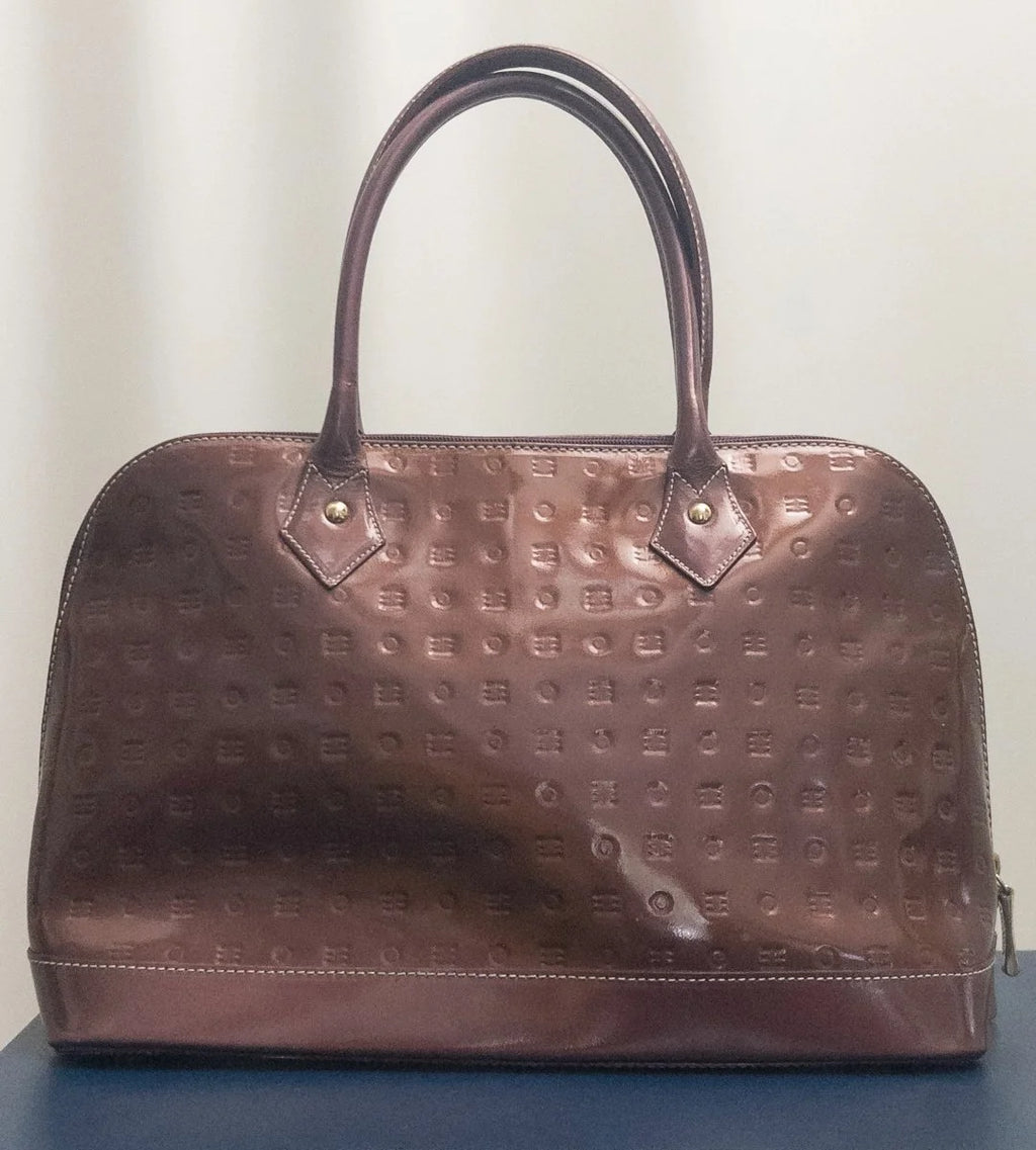 A brown embossed leather handbag with double top handles and a glossy finish, displayed on a dark blue surface against a light beige background.
