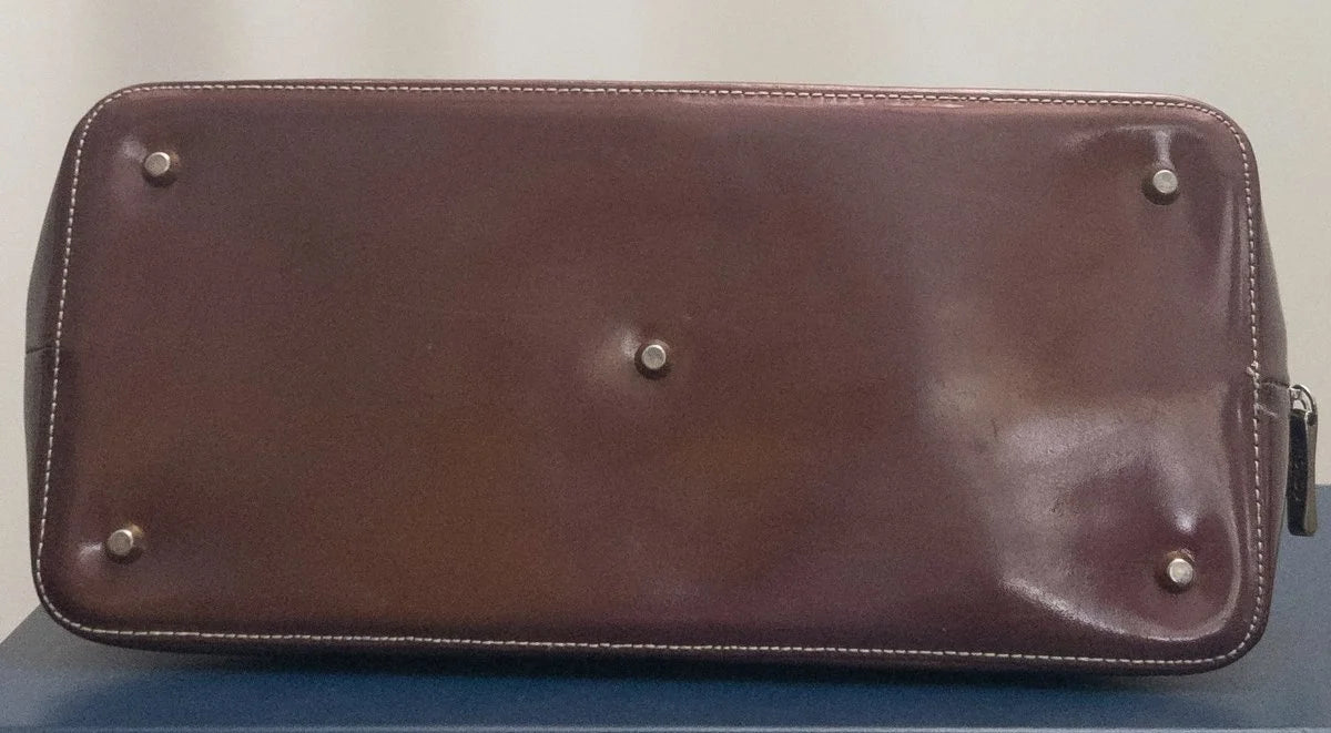 A brown leather zip-around pouch or clutch shown from the bottom, featuring silver-tone metal studs and a zipper pull on the right side.