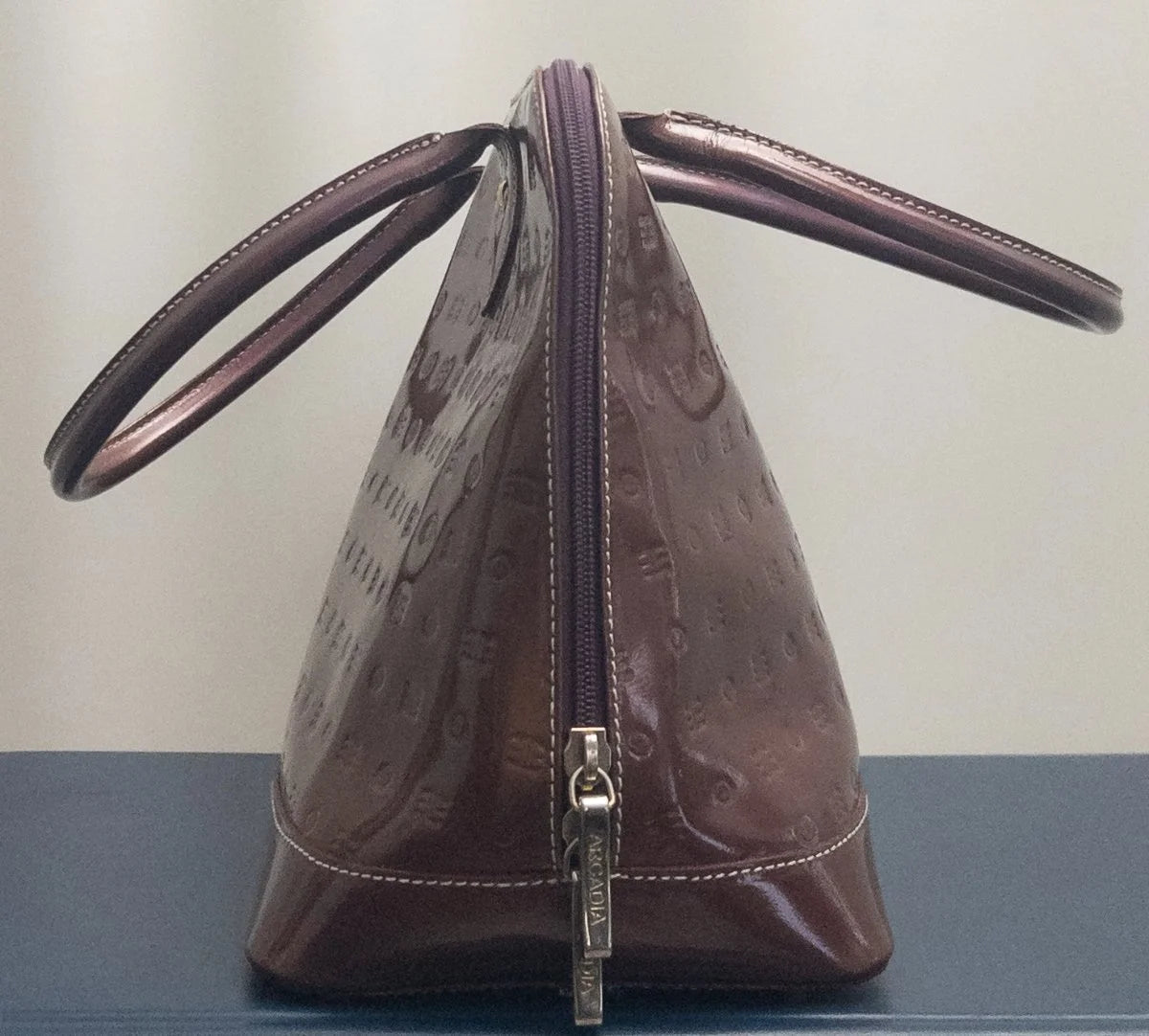 A brown Louis Vuitton shoulder bag with a glossy embossed pattern, featuring dual top handles and a front zipper closure, displayed on a dark surface against a light background.