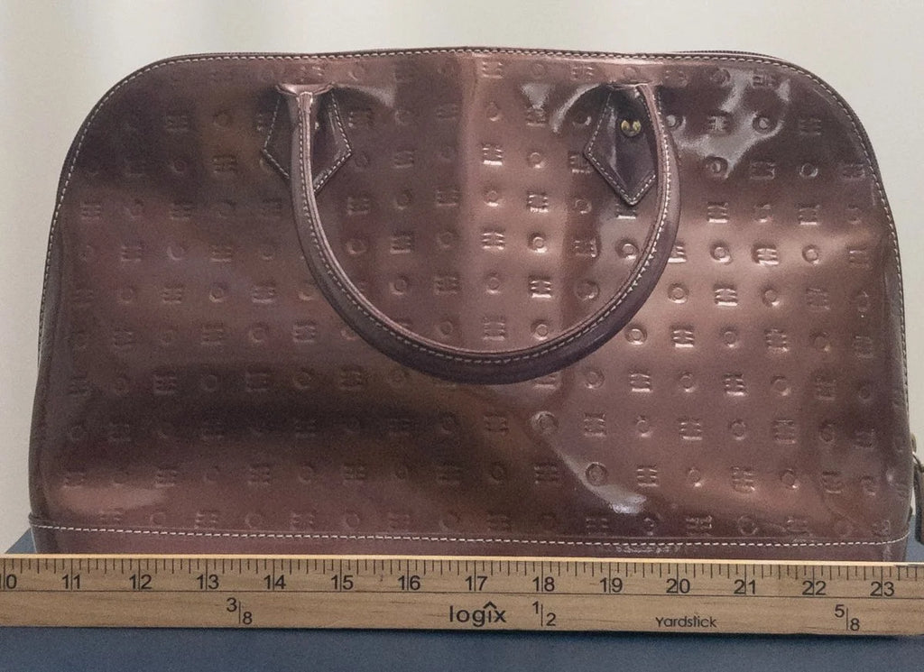 A brown embossed patent leather handbag with a curved dome shape and top handles, shown with a wooden yardstick ruler at the bottom for scale.