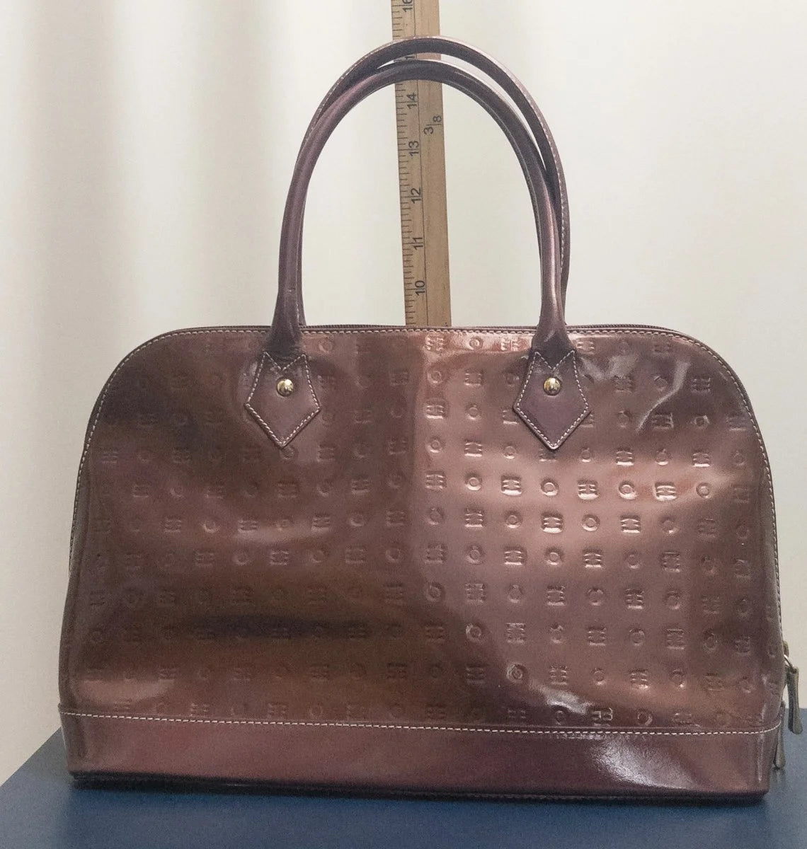 A brown patent leather handbag with an embossed monogram pattern, featuring dual top handles and a zippered closure, shown standing on a dark blue surface with a wooden ruler behind it.