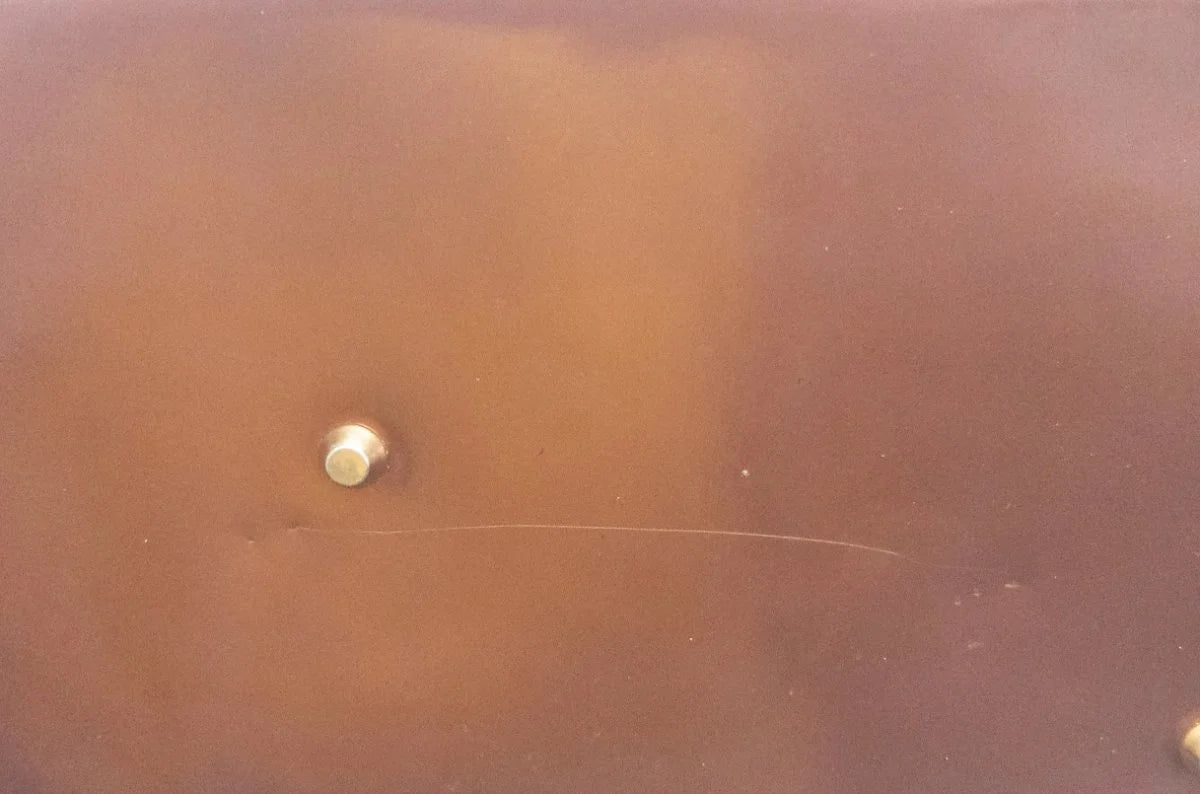 A close-up of a brown leather or faux-leather surface with a small metallic snap button and a thin scratch on the lower middle-left area.