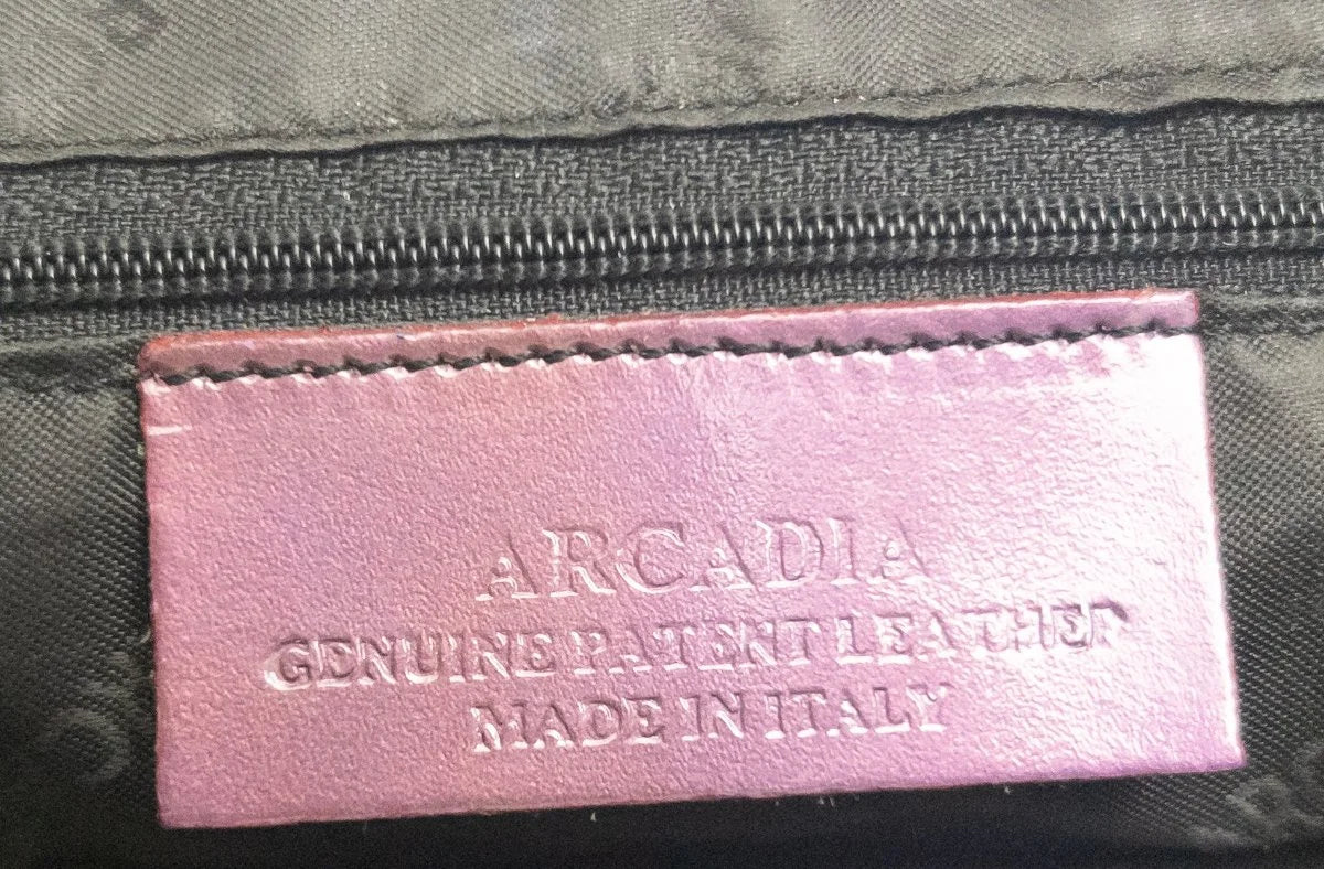 Close-up of a pink leather interior label with embossed text reading 'ARCADIA GENUINE PATENT LEATHER MADE IN ITALY' on a dark gray fabric background.
