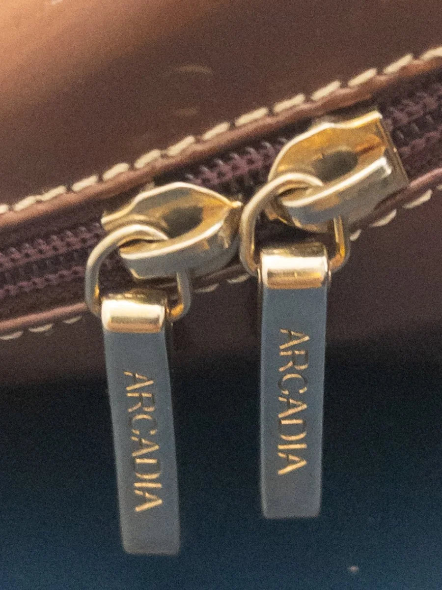 Close-up of two matching zipper pulls on a brown leather item, each with a dark gray tag printed vertically with the brand name 'ARCADIA' in gold lettering.